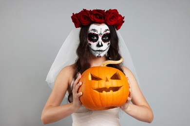 Photo of Young woman in scary bride costume with sugar skull makeup, flower crown and carved pumpkin on light grey background. Halloween celebration
