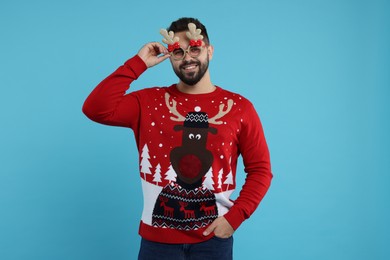 Happy young man in Christmas sweater and funny glasses on light blue background