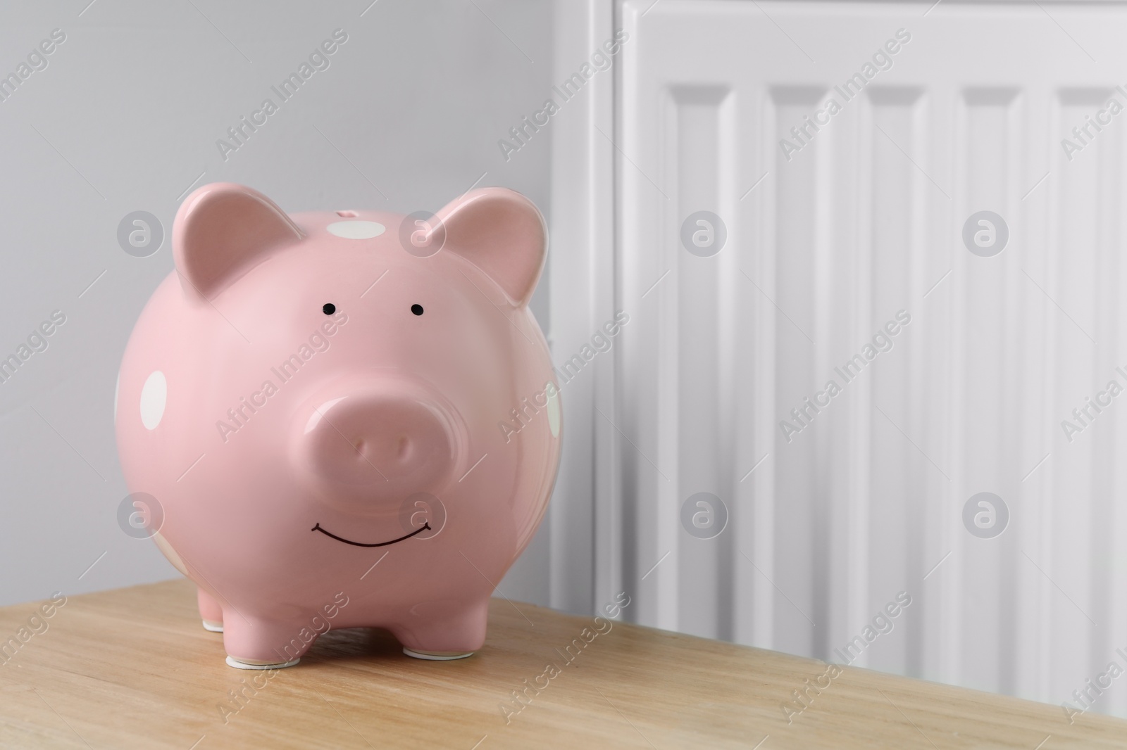 Photo of Piggy bank on wooden table near heating radiator, space for text