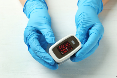 Doctor in latex gloves holding fingertip pulse oximeter at white wooden table, closeup
