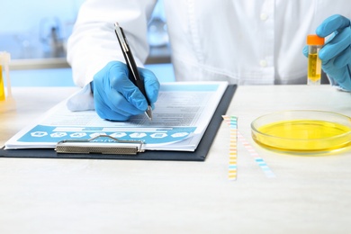 Photo of Laboratory assistant with urine sample for analysis writing results of tests at table indoors, closeup