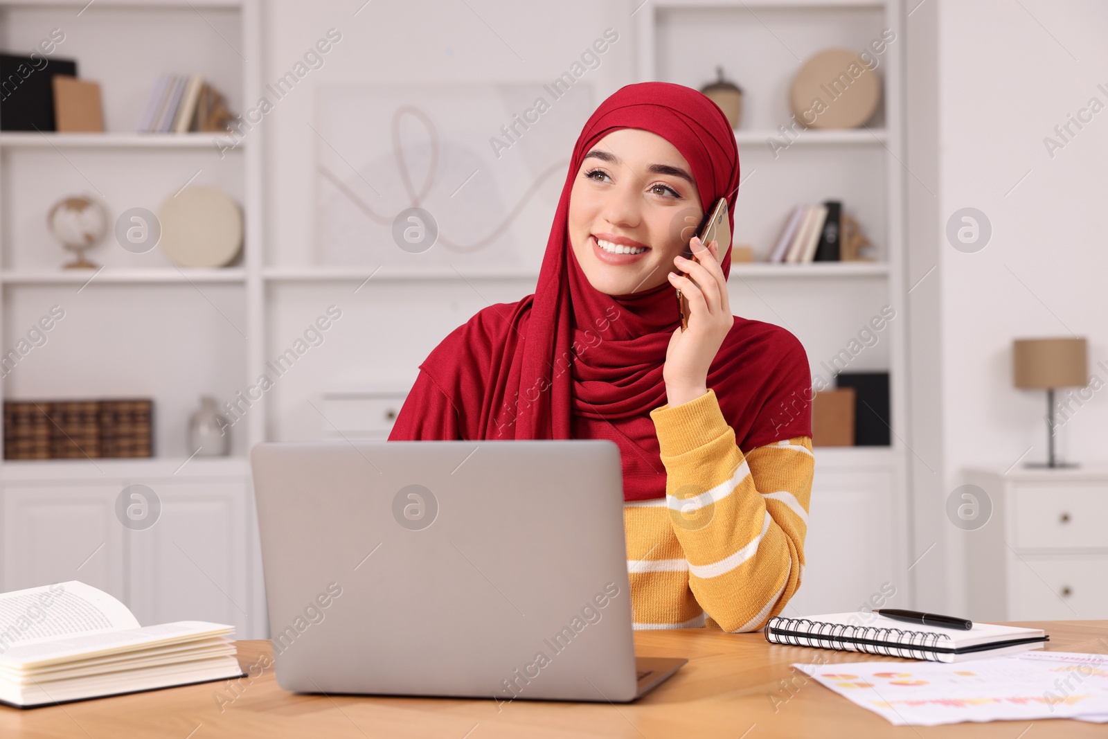 Photo of Muslim woman in hijab talking on smartphone while using laptop at wooden table in room