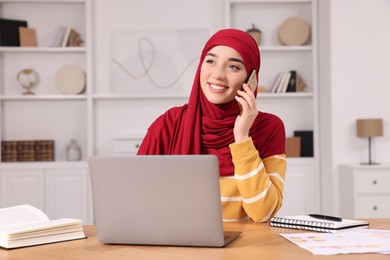 Muslim woman in hijab talking on smartphone while using laptop at wooden table in room
