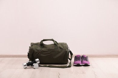 Photo of Green sports bag, sneakers and dumbbells on floor near white wall, space for text