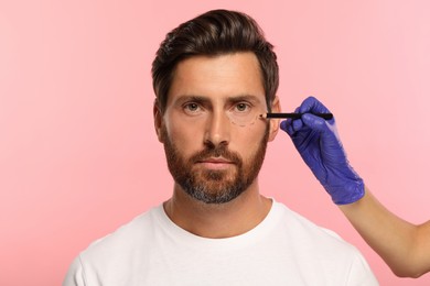 Doctor with pencil preparing patient for cosmetic surgery operation on pink background
