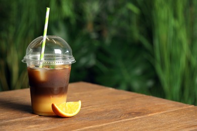 Tasty refreshing drink with coffee and orange juice in plastic cup on wooden table against blurred background, space for text