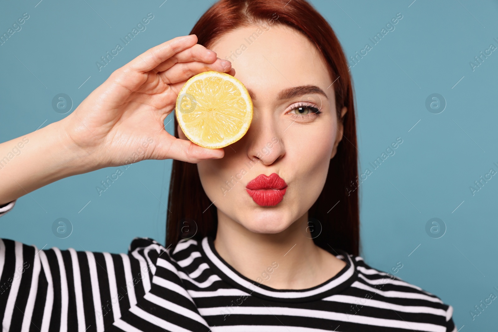 Photo of Beautiful redhead woman covering eye with lemon and blowing kiss on light blue background