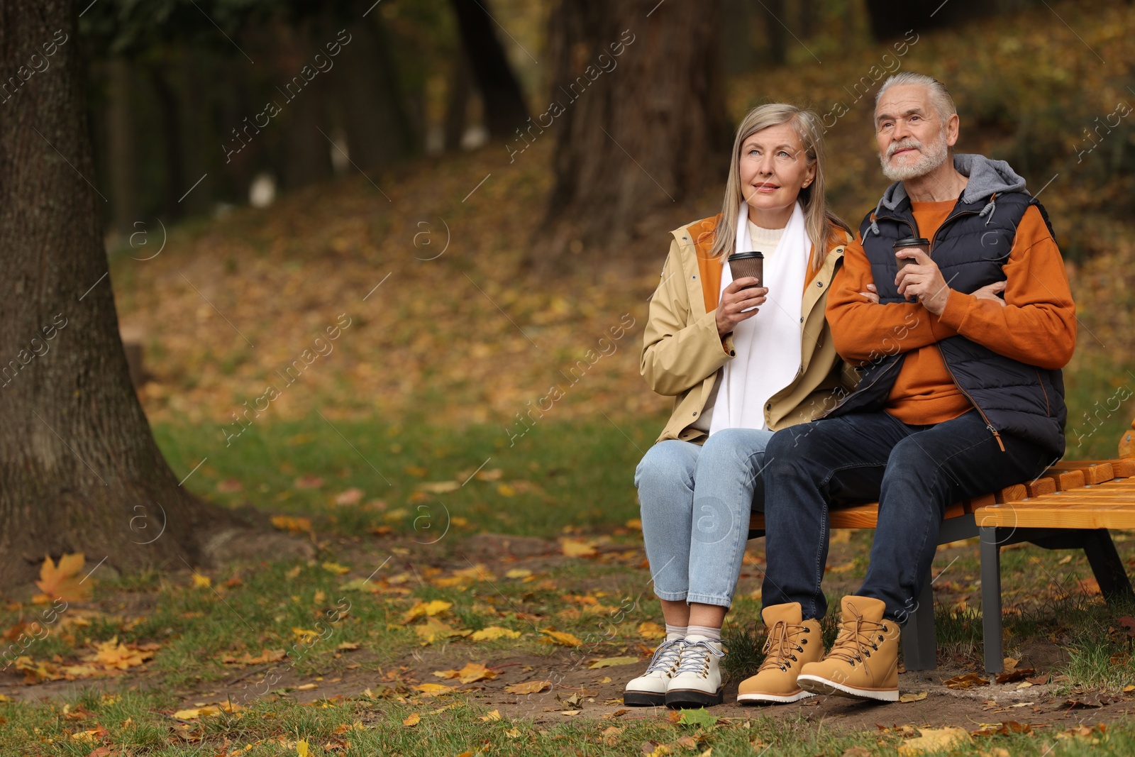 Photo of Affectionate senior couple with cups of coffee on wooden bench in autumn park, space for text