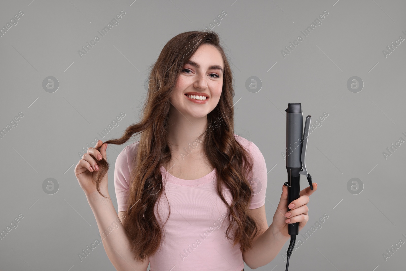 Photo of Happy young woman with beautiful hair holding curling iron on grey background
