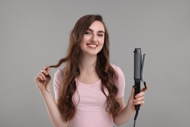 Happy young woman with beautiful hair holding curling iron on grey background