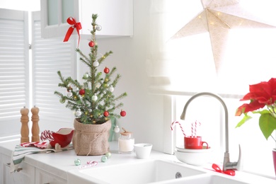 Small Christmas tree decorated with baubles and festive lights in kitchen