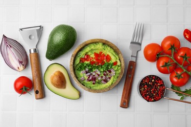Photo of Bowl of delicious guacamole with onion, tomatoes and ingredients on white tiled table, flat lay