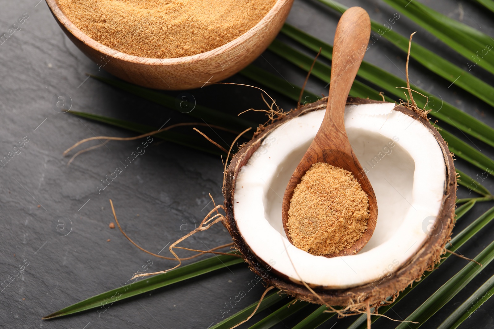 Photo of Spoon with coconut sugar, fruit, bowl and palm leaves on dark textured table, above view