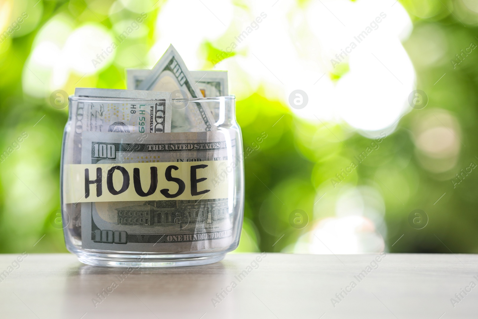 Photo of Glass jar with money and label HOUSE on table against blurred background. Space for text