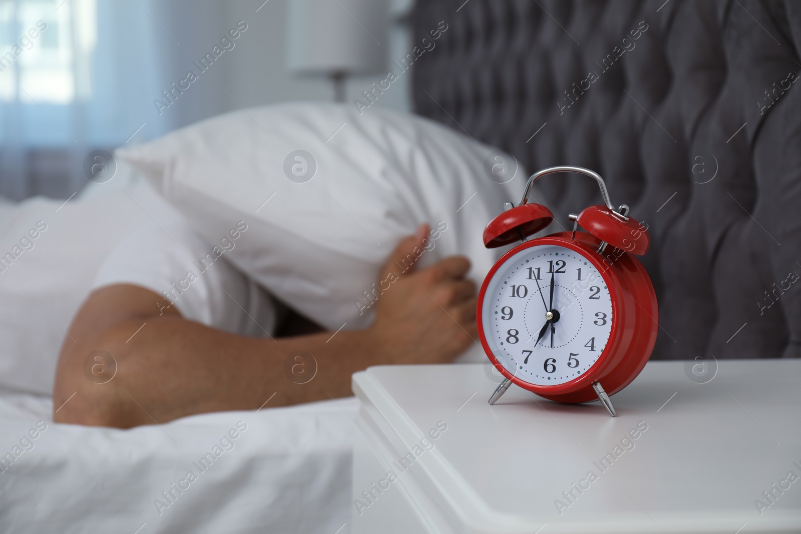 Photo of Alarm clock and man covering head with pillow in bedroom. Time of day