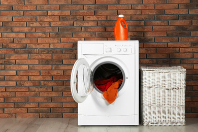 Photo of Modern washing machine with laundry, detergent and wicker basket near brick wall