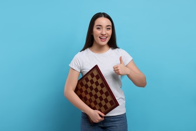 Photo of Happy woman with chessboard showing thumbs up on light blue background
