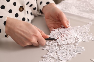 Photo of Dressmaker cutting beautiful white lace at table in atelier, closeup