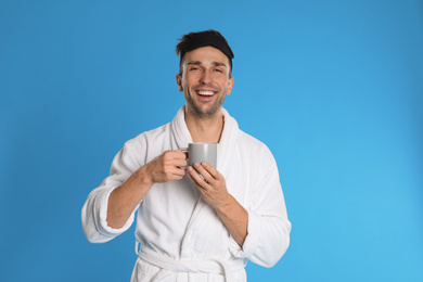 Young man in bathrobe with cup of coffee on light blue background