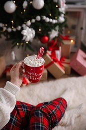 Woman with cup of delicious hot drink near Christmas tree at home, closeup