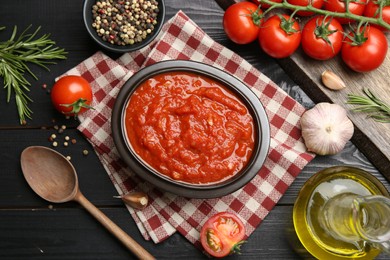 Photo of Homemade tomato sauce in bowl, spoon and fresh ingredients on black wooden table, flat lay