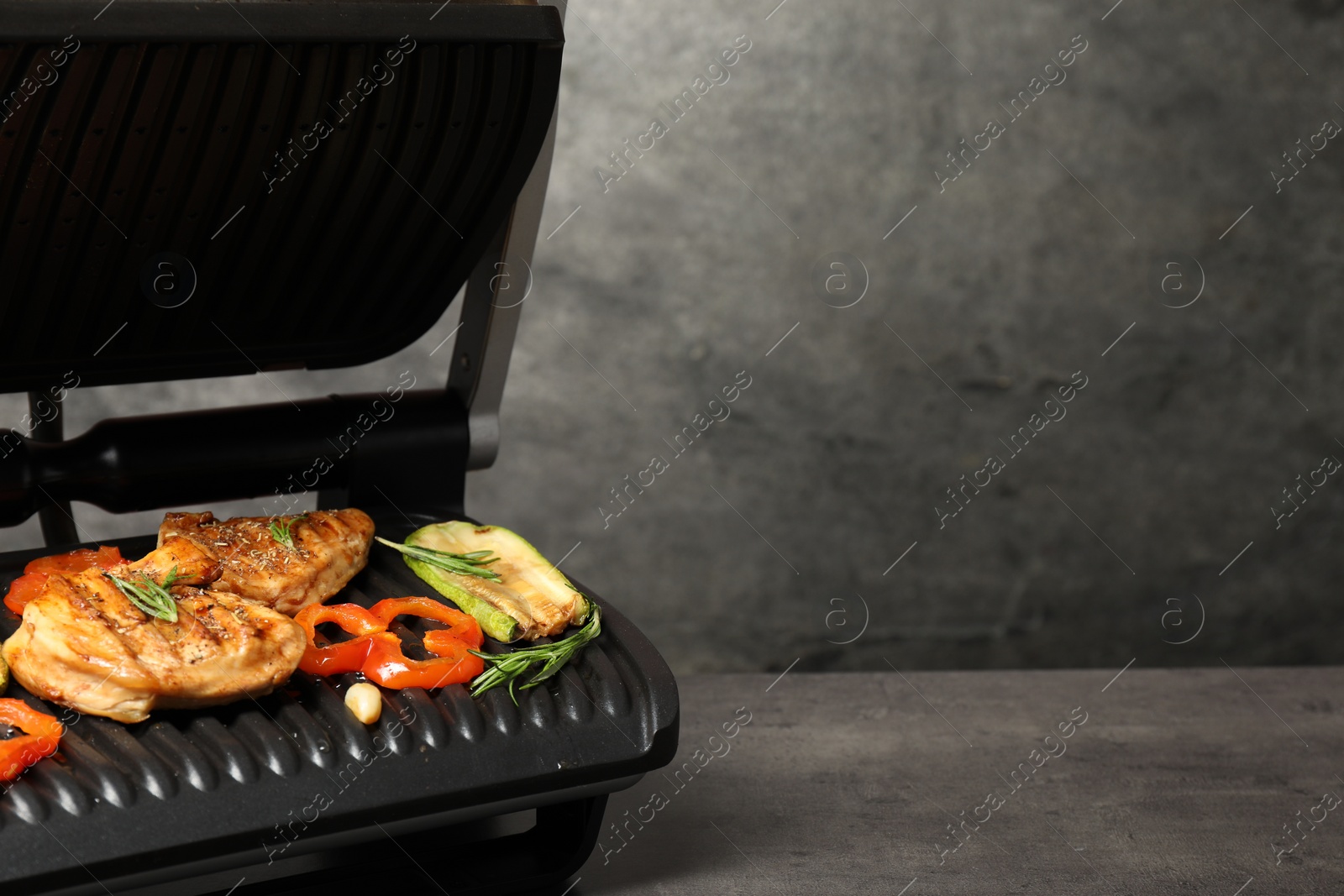 Photo of Electric grill with tasty meat, spices and vegetables on grey textured table, space for text