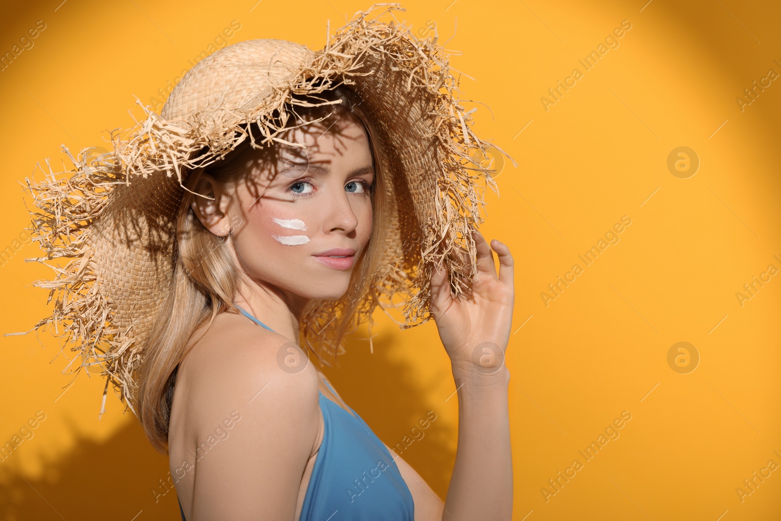 Photo of Beautiful young woman in straw hat with sun protection cream on her face against orange background, space for text