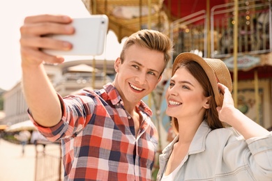 Photo of Young happy couple taking selfie near carousel in amusement park