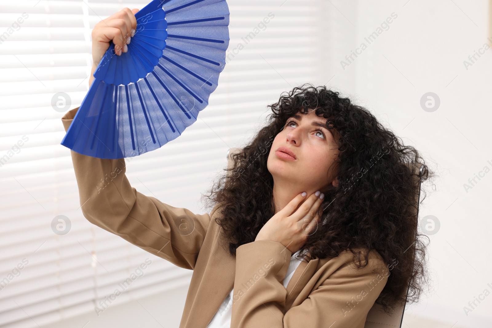 Photo of Young businesswoman waving blue hand fan to cool herself in office