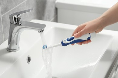 Photo of Woman washing electric toothbrush under flowing water from faucet in bathroom, closeup