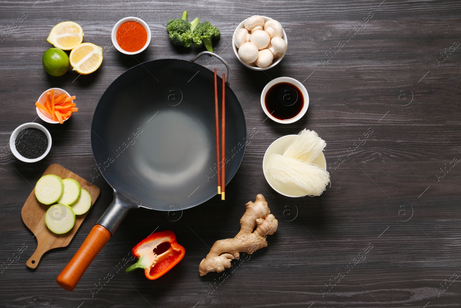 Photo of Empty iron wok and chopsticks surrounded by ingredients on dark grey wooden table, flat lay. Space for text