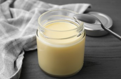 Open jar with condensed milk and spoon on dark grey wooden table, closeup