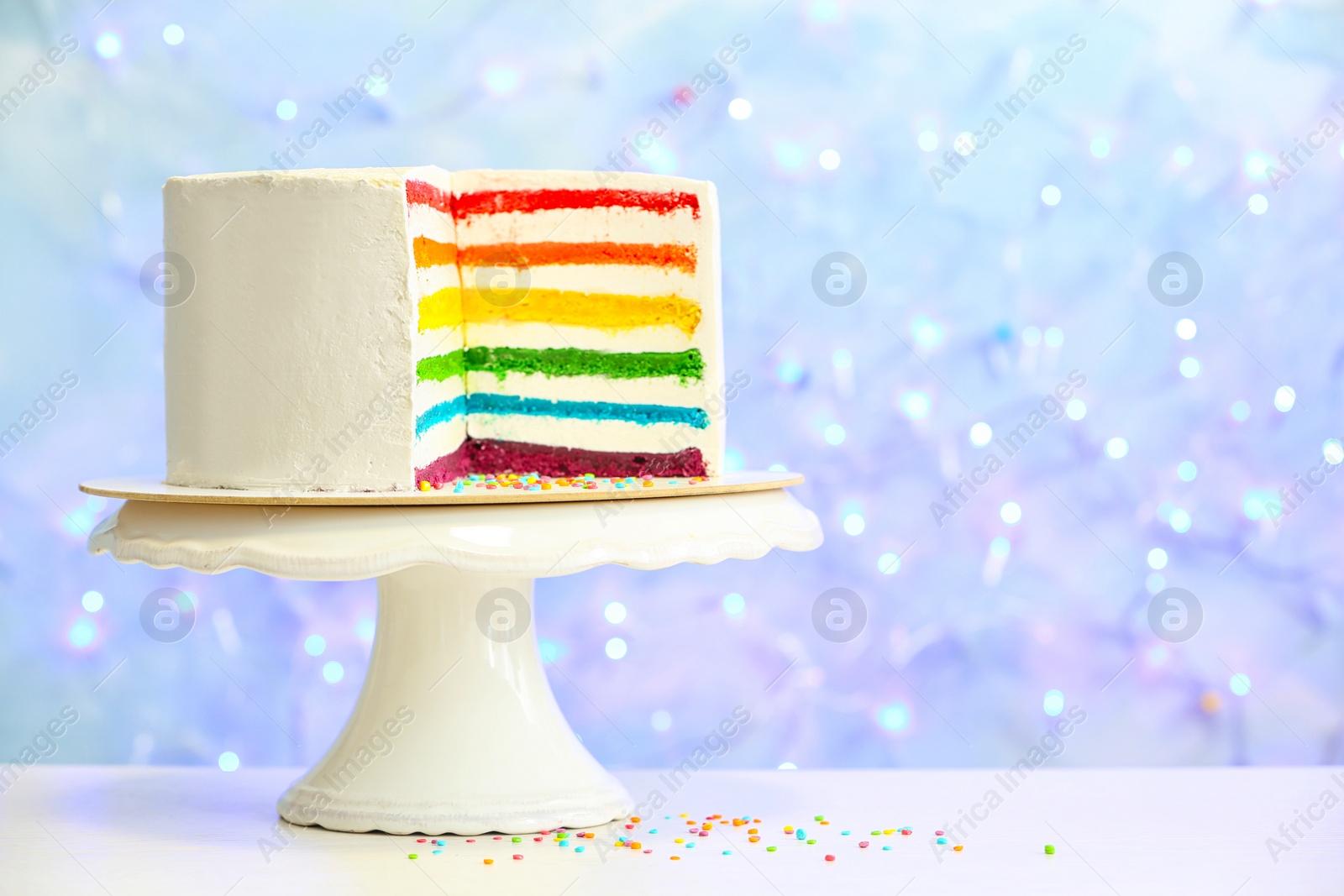 Photo of Dessert stand with delicious rainbow cake on table against blurred lights