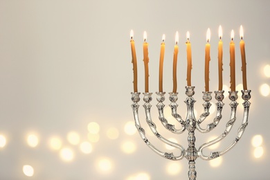 Photo of Silver menorah with burning candles against light grey background and blurred festive lights, space for text. Hanukkah celebration