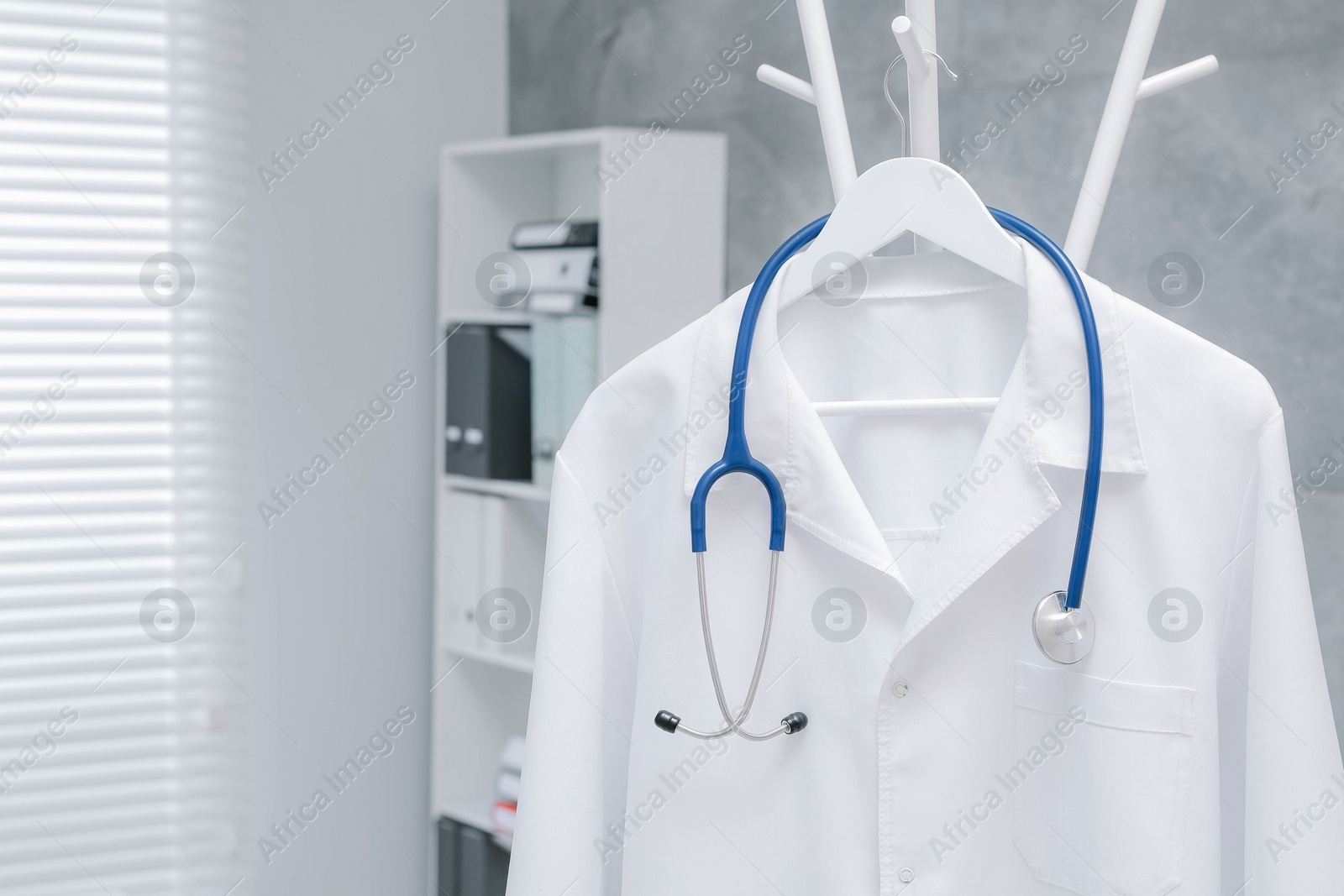 Photo of White doctor's gown and stethoscope hanging on rack in clinic, closeup. Space for text