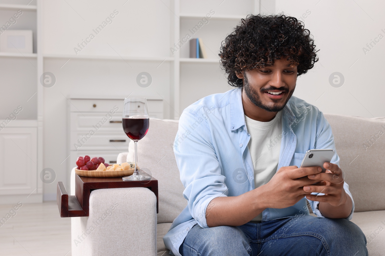 Photo of Happy man using smartphone at home. Glass of wine and snacks on sofa armrest wooden table