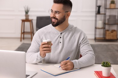Photo of Young man with cup of coffee writing in notebook at white table indoors