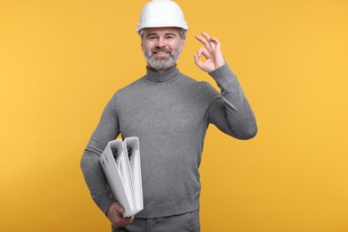 Photo of Architect in hard hat holding folders and showing ok gesture on orange background