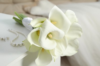 Photo of Beautiful calla lily flowers tied with ribbon and jewelry on white chest of drawers indoors, closeup