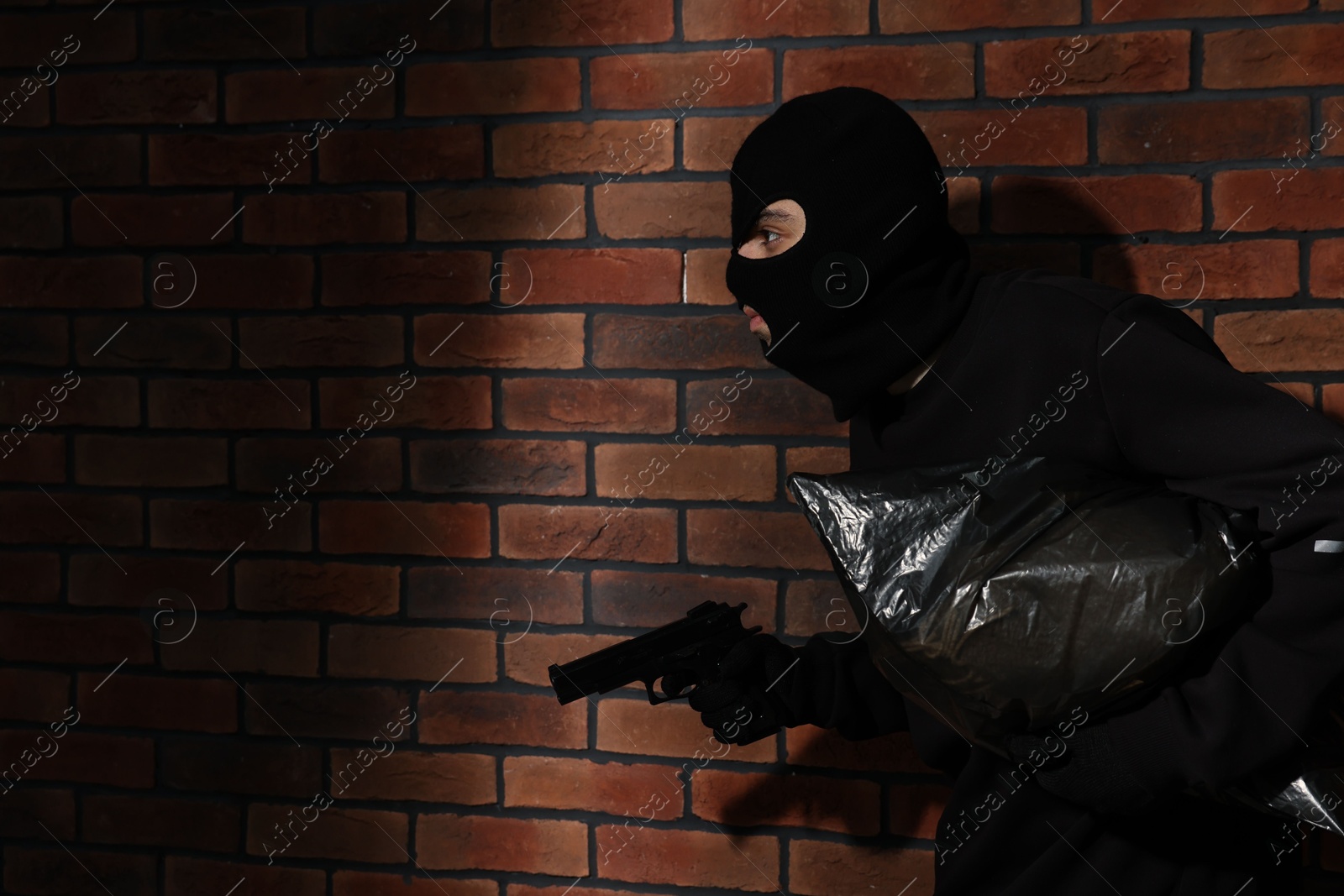 Photo of Thief in balaclava with gun and bag against red brick wall. Space for text