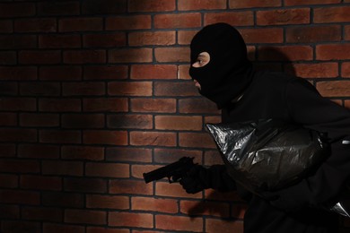 Thief in balaclava with gun and bag against red brick wall. Space for text