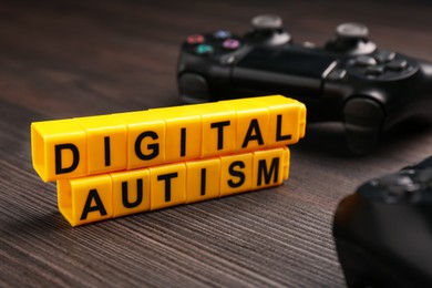 Photo of Phrase Digital Autism made of yellow cubes and gamepad on wooden table, closeup. Addictive behavior