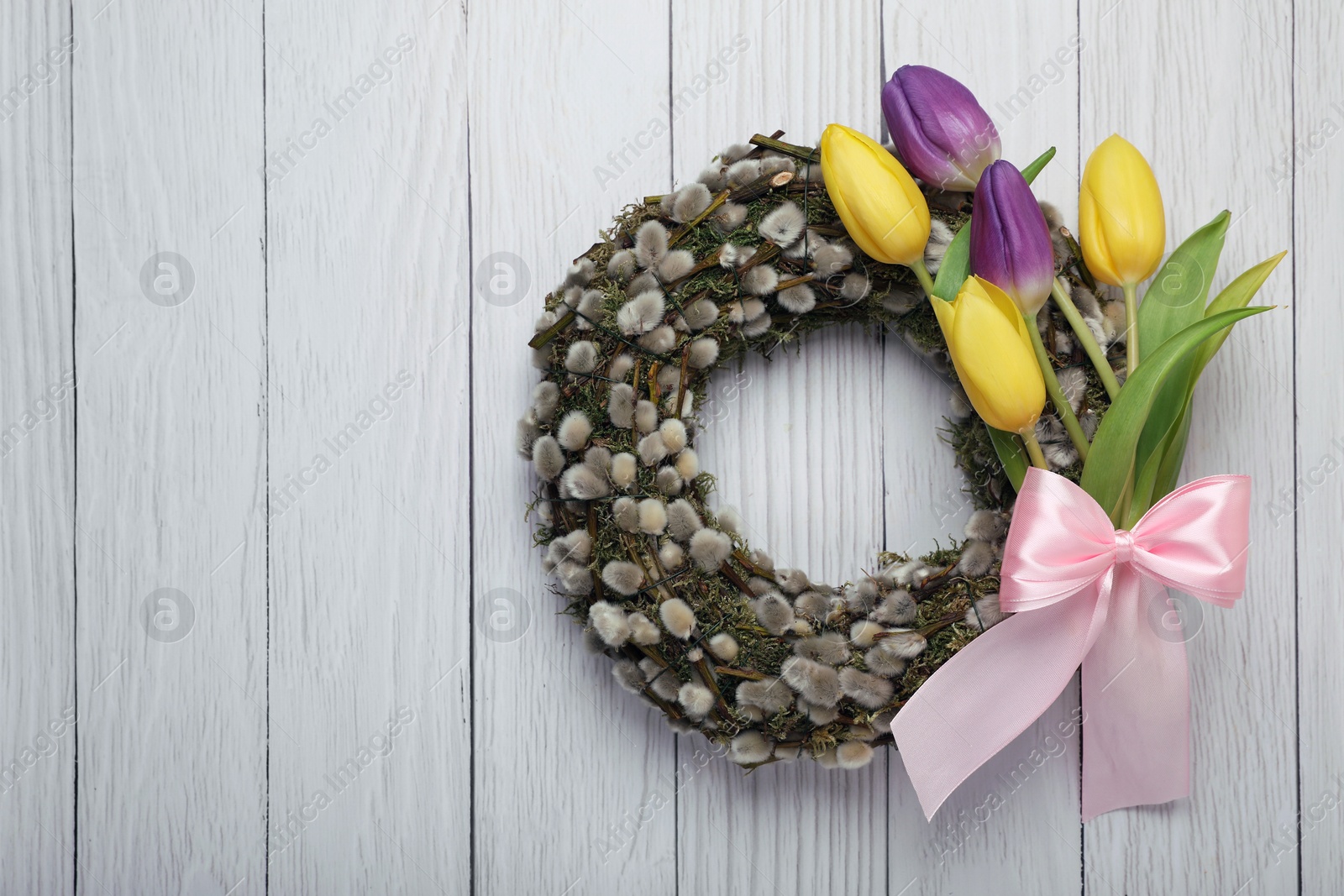 Photo of Wreath made of beautiful willow, colorful tulip flowers and pink bow on white wooden background, top view. Space for text