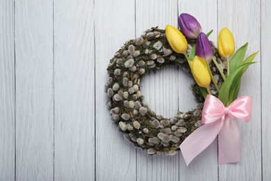 Wreath made of beautiful willow, colorful tulip flowers and pink bow on white wooden background, top view. Space for text