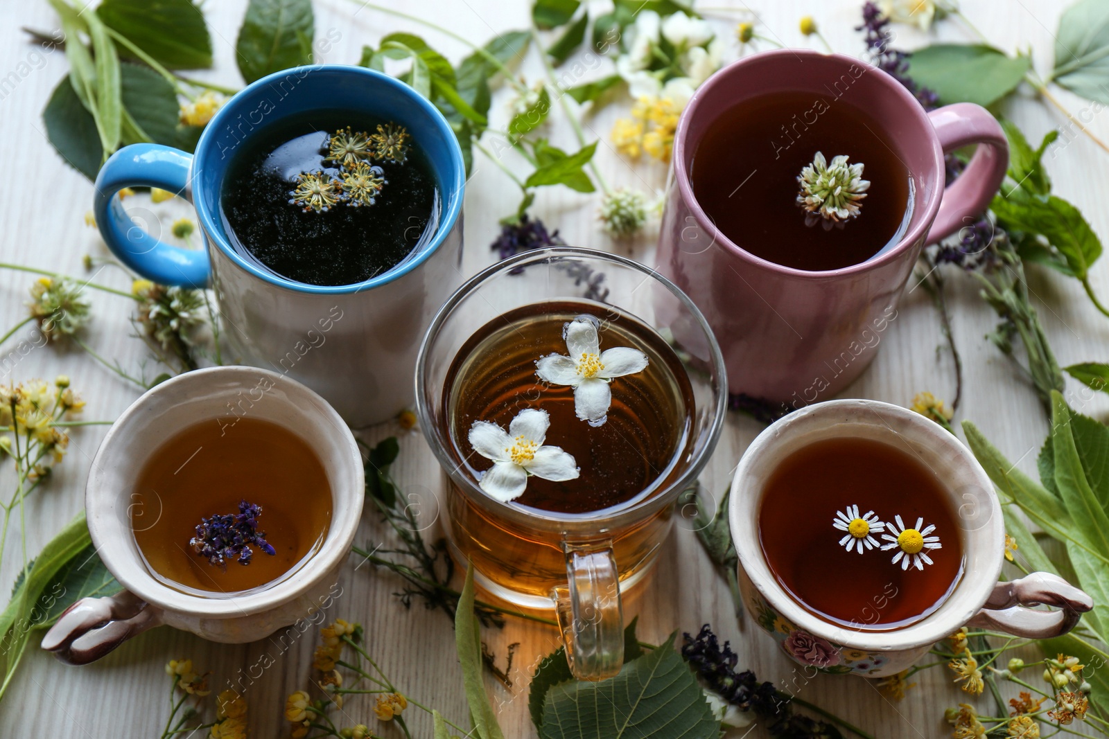 Photo of Different cups of hot aromatic tea and fresh herbs on white wooden table, above view