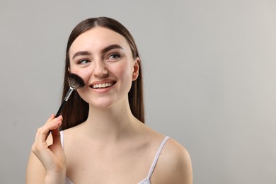 Smiling woman with freckles applying makeup with brush on grey background. Space for text