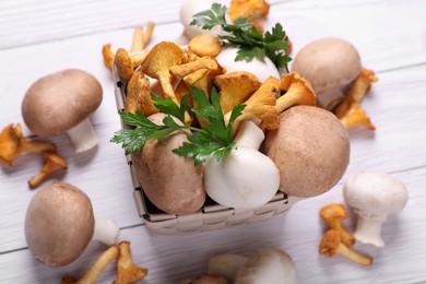Photo of Basket with different mushrooms and parsley on white wooden table, above view