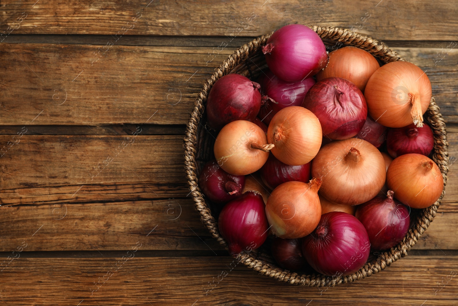 Photo of Onion bulbs in basket on wooden table, top view. Space for text