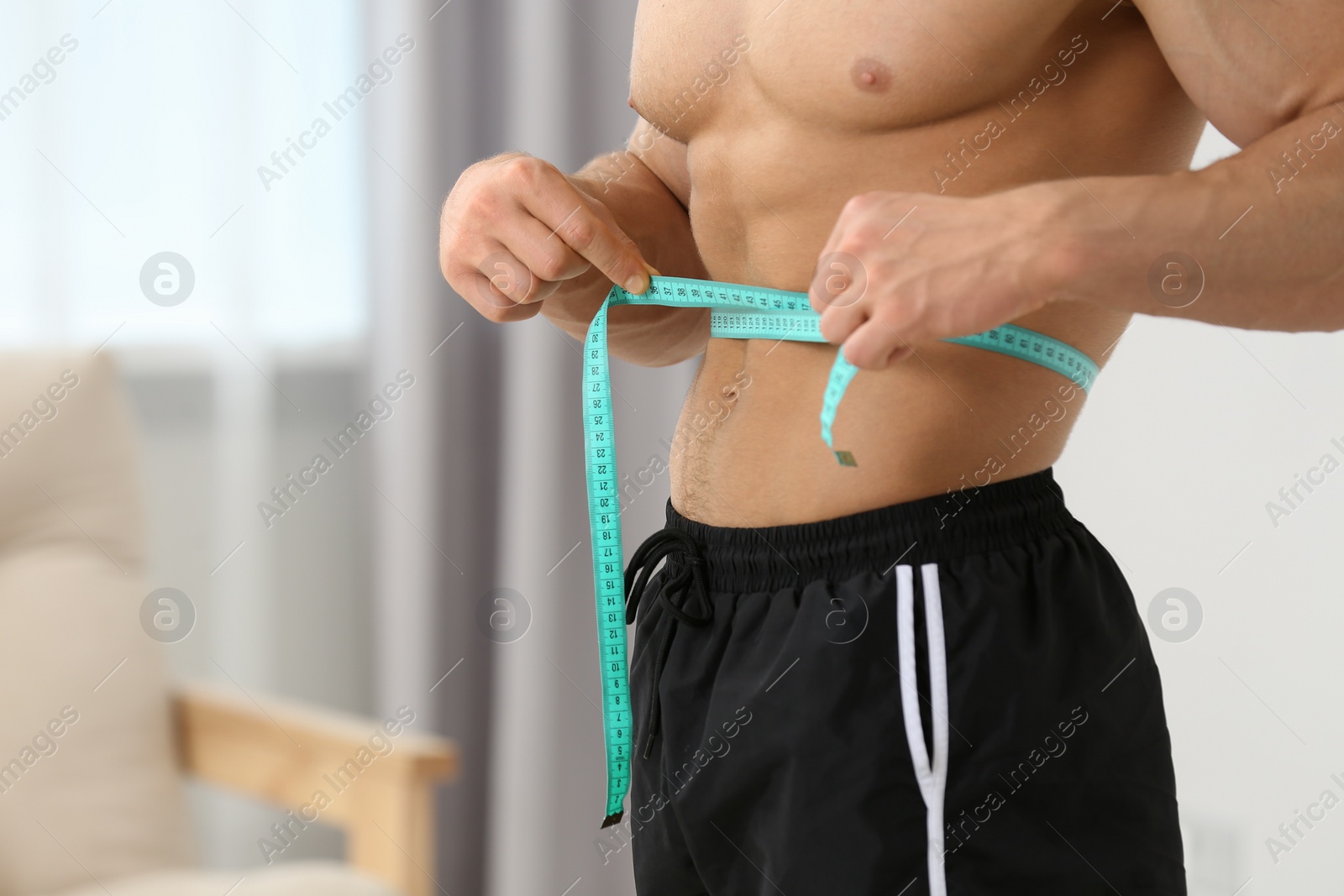 Photo of Athletic man measuring waist with tape at home, closeup and space for text. Weight loss concept