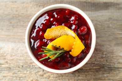 Photo of Fresh cranberry sauce in bowl, rosemary and orange peel on wooden table, top view